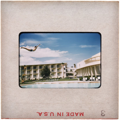 A person is diving off a pool into the water, with a large hotel building in the background.