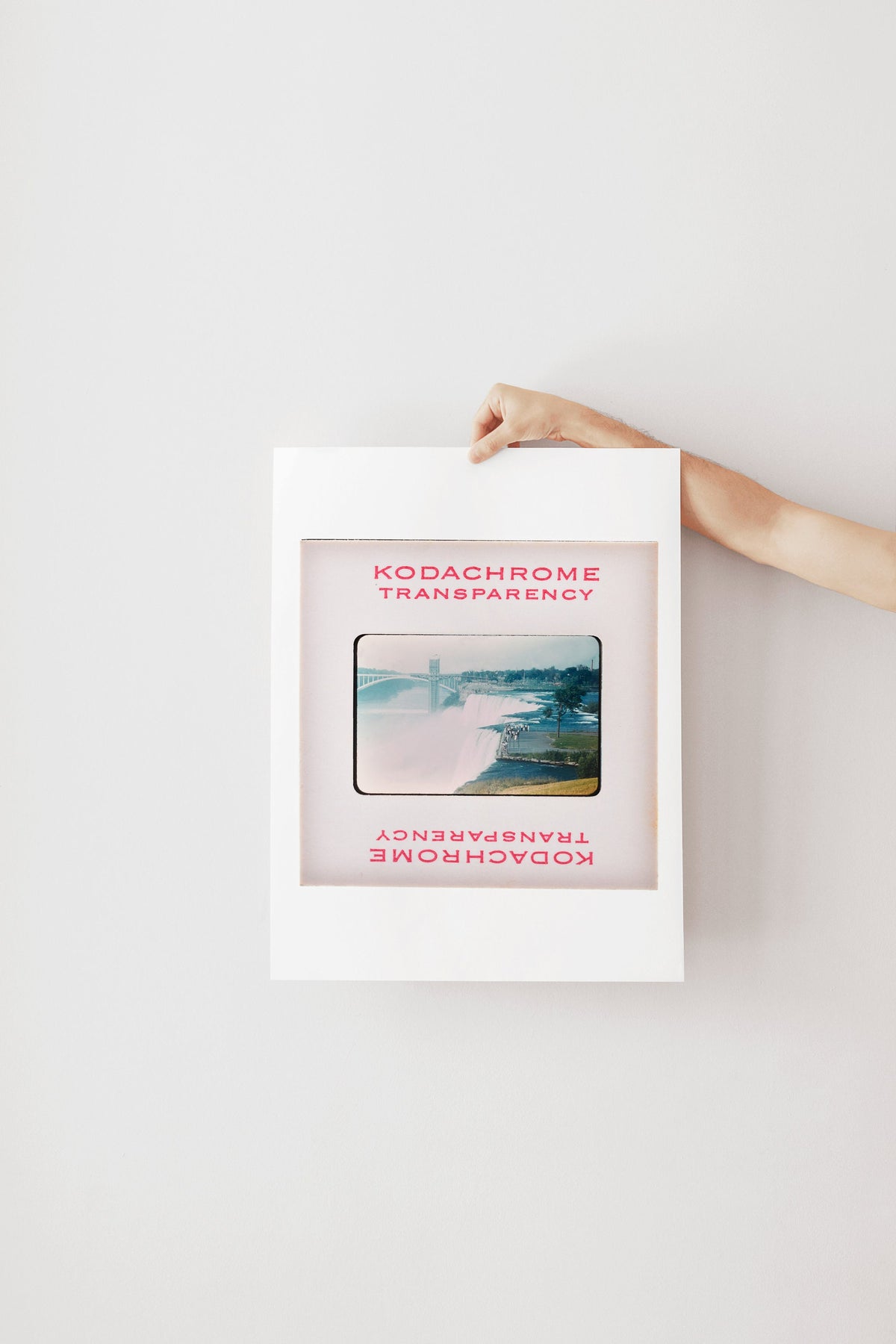 A person's hand is holding a framed photograph of a scenic landscape, with the text "Kodachrome transparency" visible on the frame.