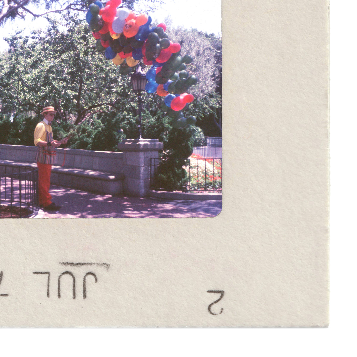 A man in a yellow shirt and orange pants is holding a bunch of colorful balloons in a park setting.