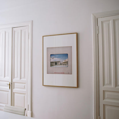 a framed picture hanging on a white wall, with a white door and a white chair visible in the background.