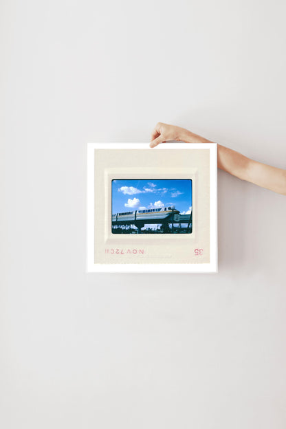 A person's hand is holding a framed picture of a train against a white wall.