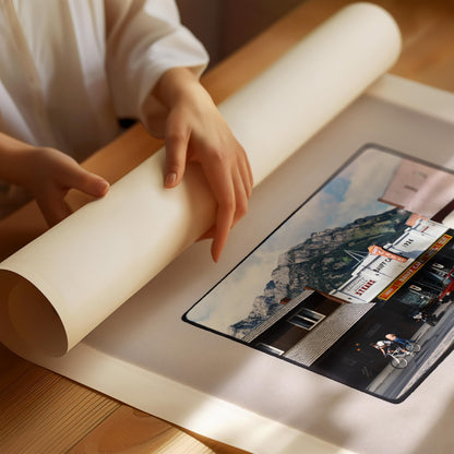 A person's hands are holding a roll of paper, which appears to be a canvas or print, over a printed image of a building and a mountain landscape.