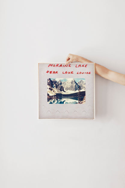A person's hand is holding a framed photograph of a scenic mountain landscape, with the text "MORaine Lake" and "Year Lake Louise" written on the frame.