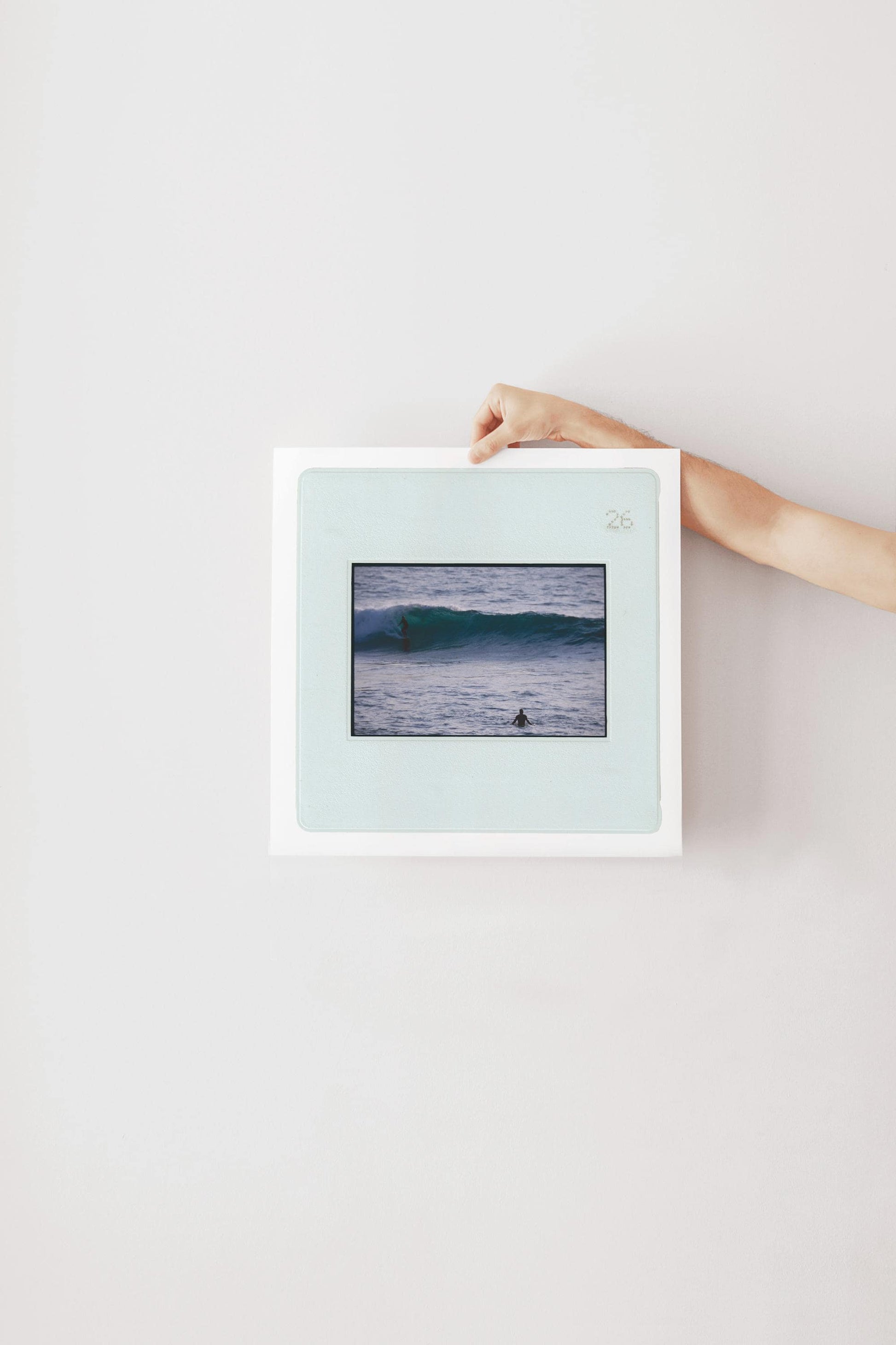 Vintage slide photograph of two surfers in the ocean — one riding a glassy teal-green wave in the background and one waiting chest-deep in silver-grey water in the foreground, shot with a telephoto lens from shore. Printed as a giclée fine art print on archival matte paper.