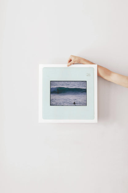 Vintage slide photograph of two surfers in the ocean — one riding a glassy teal-green wave in the background and one waiting chest-deep in silver-grey water in the foreground, shot with a telephoto lens from shore. Printed as a giclée fine art print on archival matte paper.