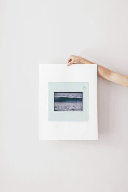 Vintage slide photograph of two surfers in the ocean — one riding a glassy teal-green wave in the background and one waiting chest-deep in silver-grey water in the foreground, shot with a telephoto lens from shore. Printed as a giclée fine art print on archival matte paper.