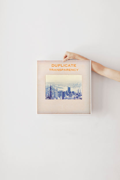 A person's hand is holding a framed picture of a cityscape, with the text "DUPLICATE TRANSPARENCY" written on the frame.