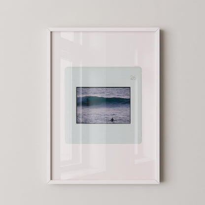 Vintage slide photograph of two surfers in the ocean — one riding a glassy teal-green wave in the background and one waiting chest-deep in silver-grey water in the foreground, shot with a telephoto lens from shore. Printed as a giclée fine art print on archival matte paper.