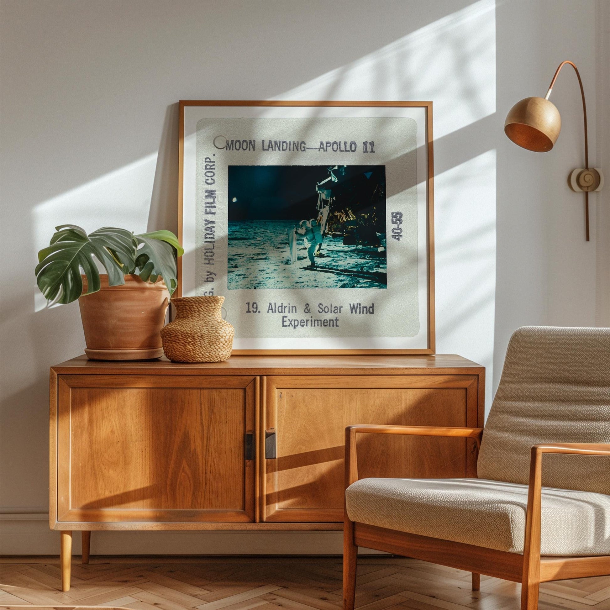 a framed photograph of a person on the moon, with the text "Moon Landing - Apollo 11" and "40.55" visible. The photograph is displayed on a wooden cabinet, with a potted plant and a woven basket on top. A beige armchair is positioned next to the cabinet, and a lamp is visible in the background.