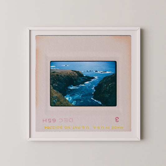 The image is a framed photograph of a scenic coastal landscape with rocky cliffs, crashing waves, and a vast expanse of blue ocean.