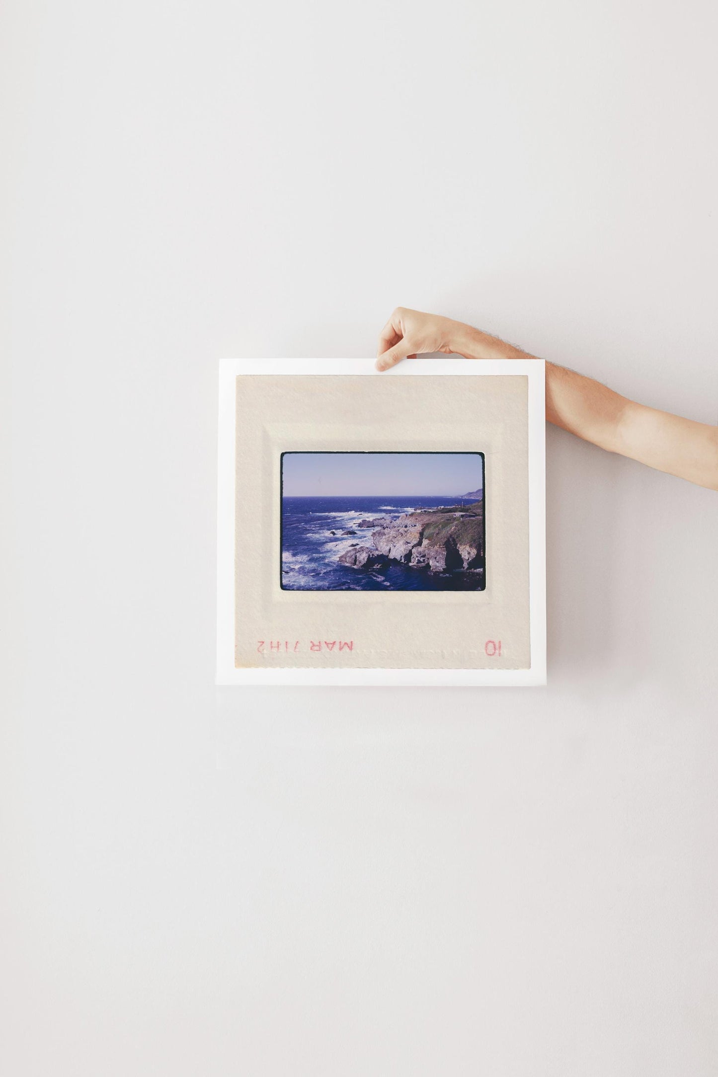Vintage 1971 Kodachrome slide photograph of the Carmel California coastline, showing dramatic rocky cliffs and outcroppings meeting deep cobalt Pacific Ocean waters, with white surf, a green headland, and a small structure visible at the cliff's edge. Printed as a giclée fine art print on archival matte paper.
