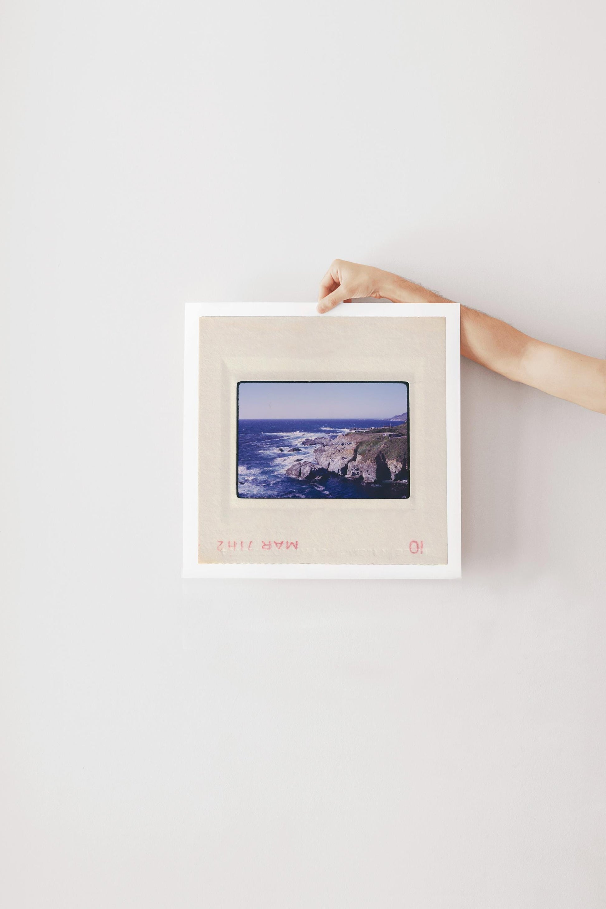 Vintage 1971 Kodachrome slide photograph of the Carmel California coastline, showing dramatic rocky cliffs and outcroppings meeting deep cobalt Pacific Ocean waters, with white surf, a green headland, and a small structure visible at the cliff's edge. Printed as a giclée fine art print on archival matte paper.