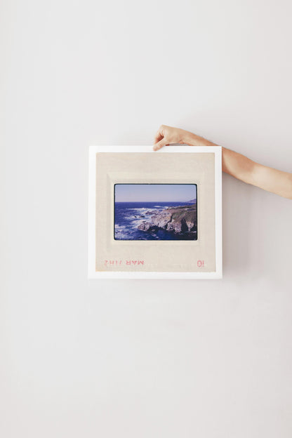 Vintage 1971 Kodachrome slide photograph of the Carmel California coastline, showing dramatic rocky cliffs and outcroppings meeting deep cobalt Pacific Ocean waters, with white surf, a green headland, and a small structure visible at the cliff's edge. Printed as a giclée fine art print on archival matte paper.