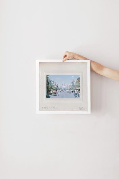 A person's hand is holding a white picture frame with a framed photograph of a city street scene inside it.