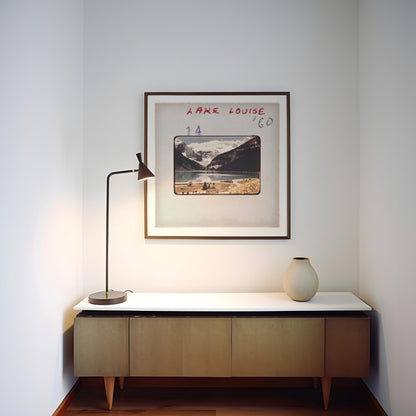 a framed picture of a mountain landscape hanging on a wall above a wooden cabinet with a white countertop. There is a lamp on the cabinet and a vase on the countertop.