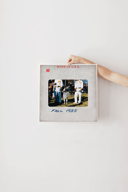 A person's hand is holding a framed photograph of two people in baseball uniforms, with the text "FALL 1955" printed on the frame.