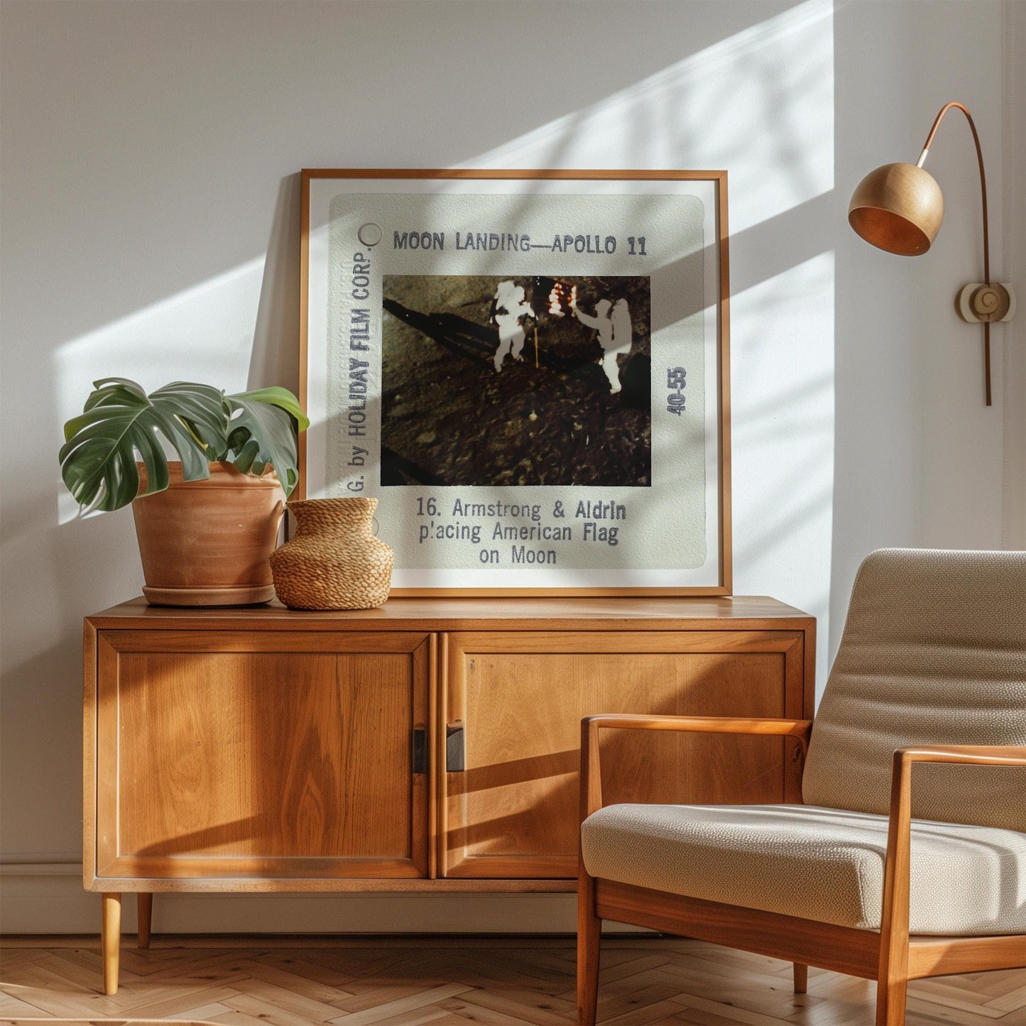 a framed photograph of the Apollo 11 moon landing, with the text "Moon Landing - Apollo 11" and "16. Armstrong & Aldrin Placing American Flag on Moon" visible. The photograph is displayed on a wooden cabinet, with a potted plant and a woven basket on top. A beige armchair is positioned in the foreground, and a lamp is visible in the background.
