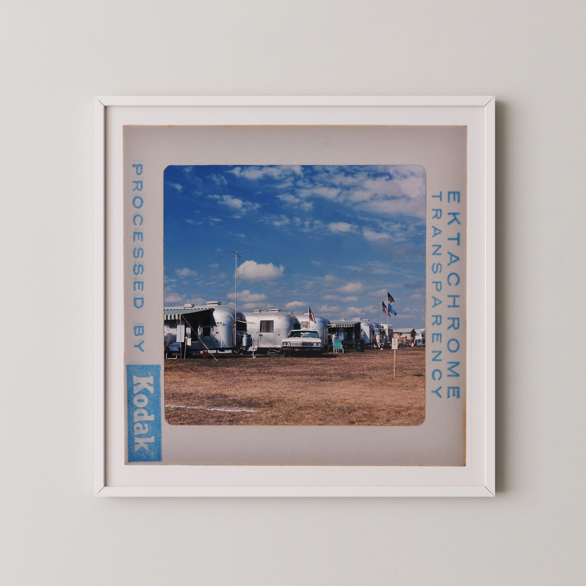 A framed photograph of a trailer park with various trailers and people in the background, set against a blue sky with clouds.