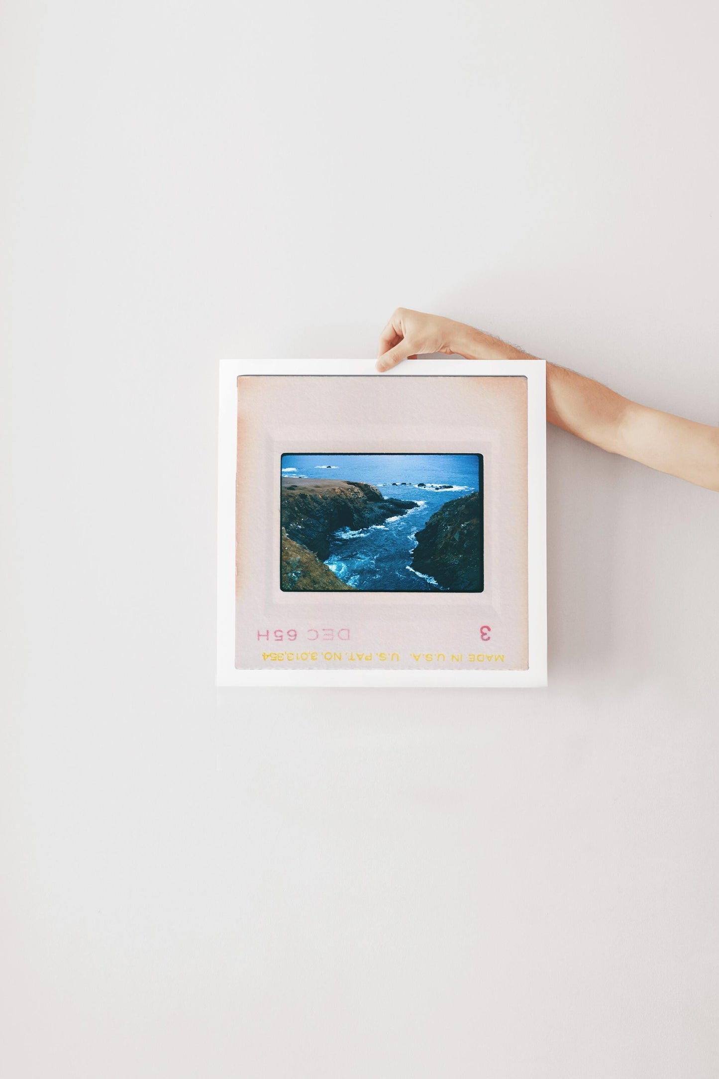 Vintage 1965 Kodachrome slide photograph taken from an elevated vantage point looking down into a narrow rocky inlet along the California coast, with deep teal water churning between two rugged headlands and scattered rocks visible in the open Pacific Ocean beyond. Printed as a giclée fine art print on archival matte paper.