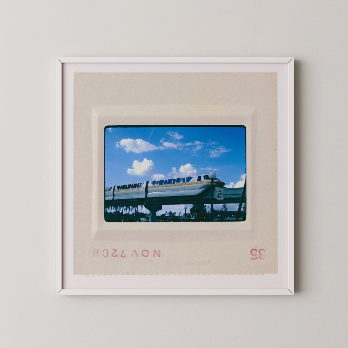 A framed photograph of a monorail train on a track against a blue sky with clouds.