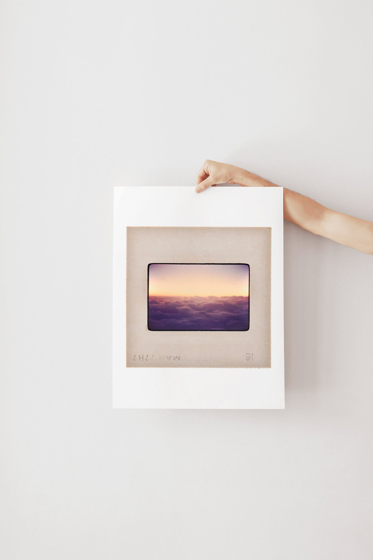 A hand is holding a framed picture of a sunset over a body of water, with the frame positioned on a white wall.