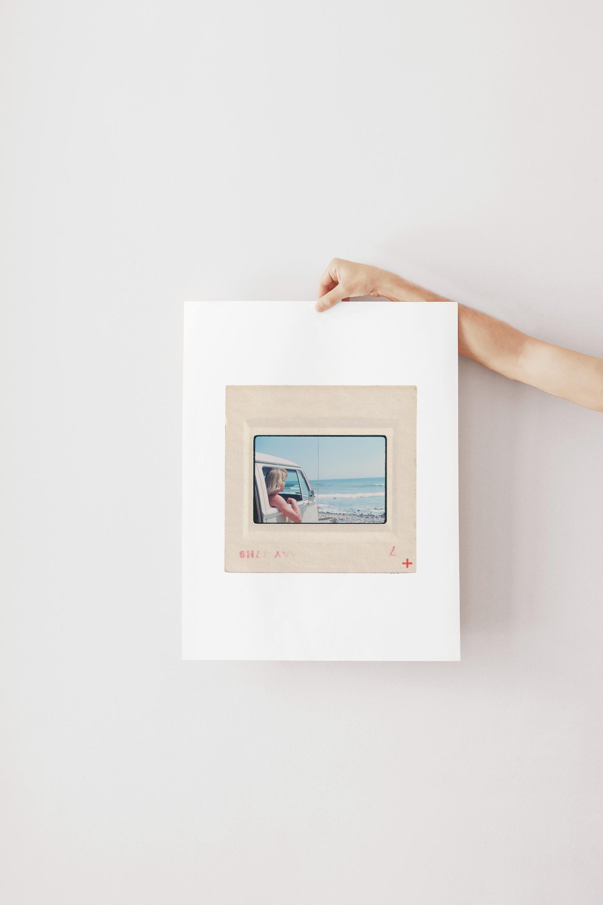 Vintage 1977 Kodachrome slide photograph of a blonde child leaning out the window of a white Volkswagen Microbus, looking toward the ocean along the California coast. Rocky shoreline and gentle surf visible in the background. Printed as a giclée fine art print on archival matte paper.