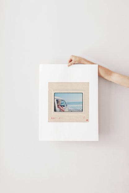 Vintage 1977 Kodachrome slide photograph of a blonde child leaning out the window of a white Volkswagen Microbus, looking toward the ocean along the California coast. Rocky shoreline and gentle surf visible in the background. Printed as a giclée fine art print on archival matte paper.