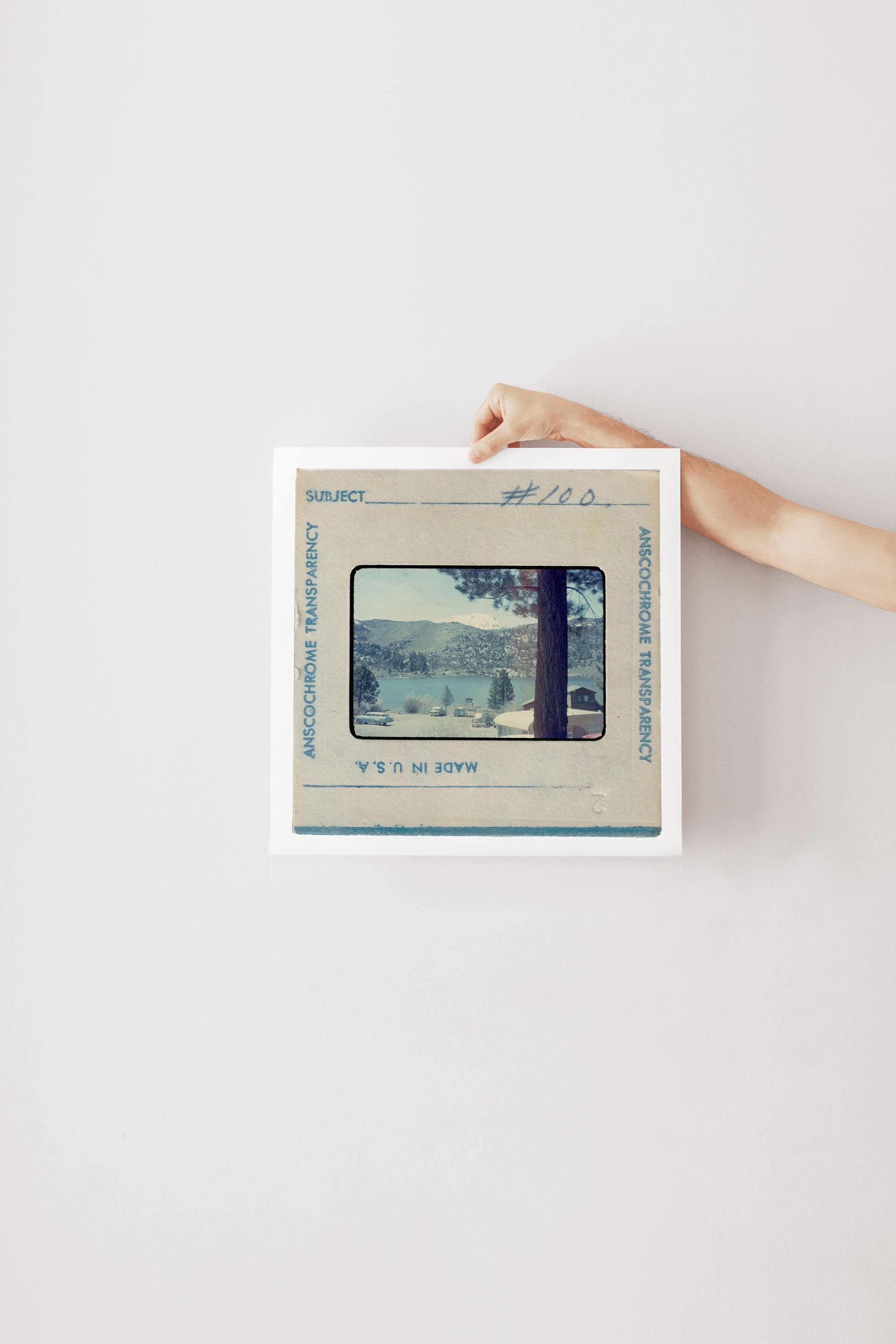Vintage Anscochrome slide photograph of a mountain lake campground community, showing pine-forested hills, a snow-capped peak, classic cars parked along the shoreline, and lakeside cabins, viewed from beneath a tall pine tree. Printed as a giclée fine art print on archival matte paper.