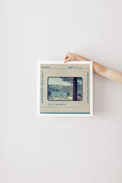 Vintage Anscochrome slide photograph of a mountain lake campground community, showing pine-forested hills, a snow-capped peak, classic cars parked along the shoreline, and lakeside cabins, viewed from beneath a tall pine tree. Printed as a giclée fine art print on archival matte paper.