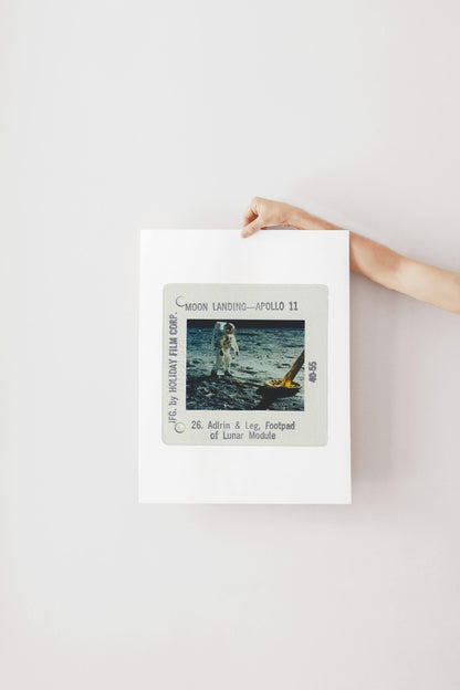 A person's hand is holding a framed photograph of the Apollo 11 moon landing, featuring the astronaut Adrin and the lunar module.