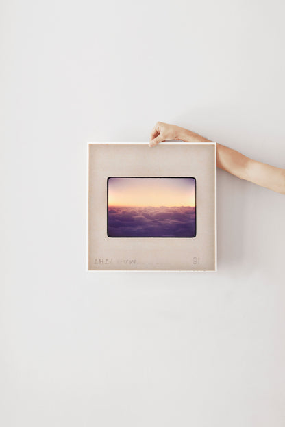 A person's hand is holding a framed picture of a sunset over a body of water.