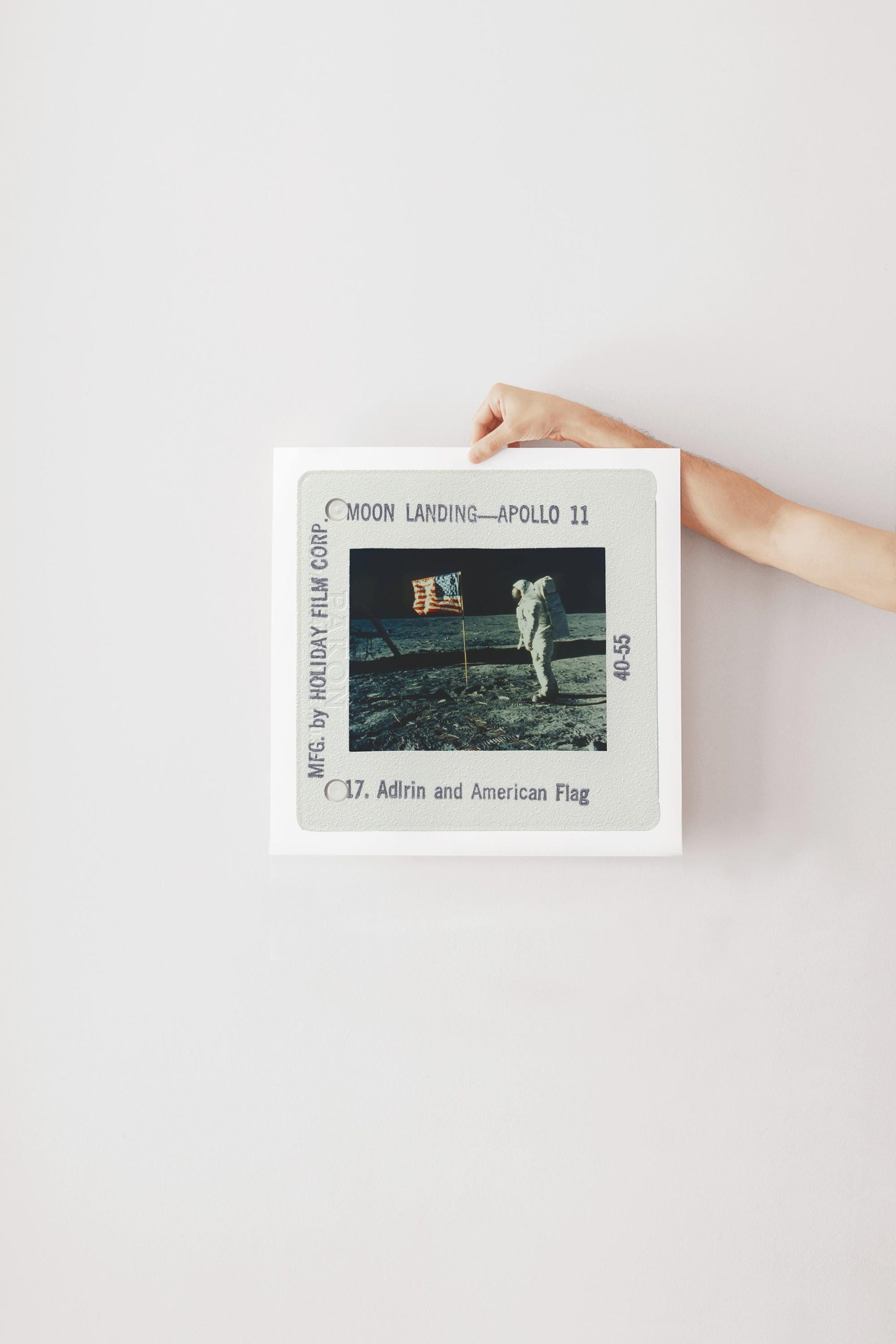 A person's hand is holding a framed photograph of the Apollo 11 moon landing, with the caption "Moon Landing - Apollo 11" and the date "C.17. Adrin and American Flag".
