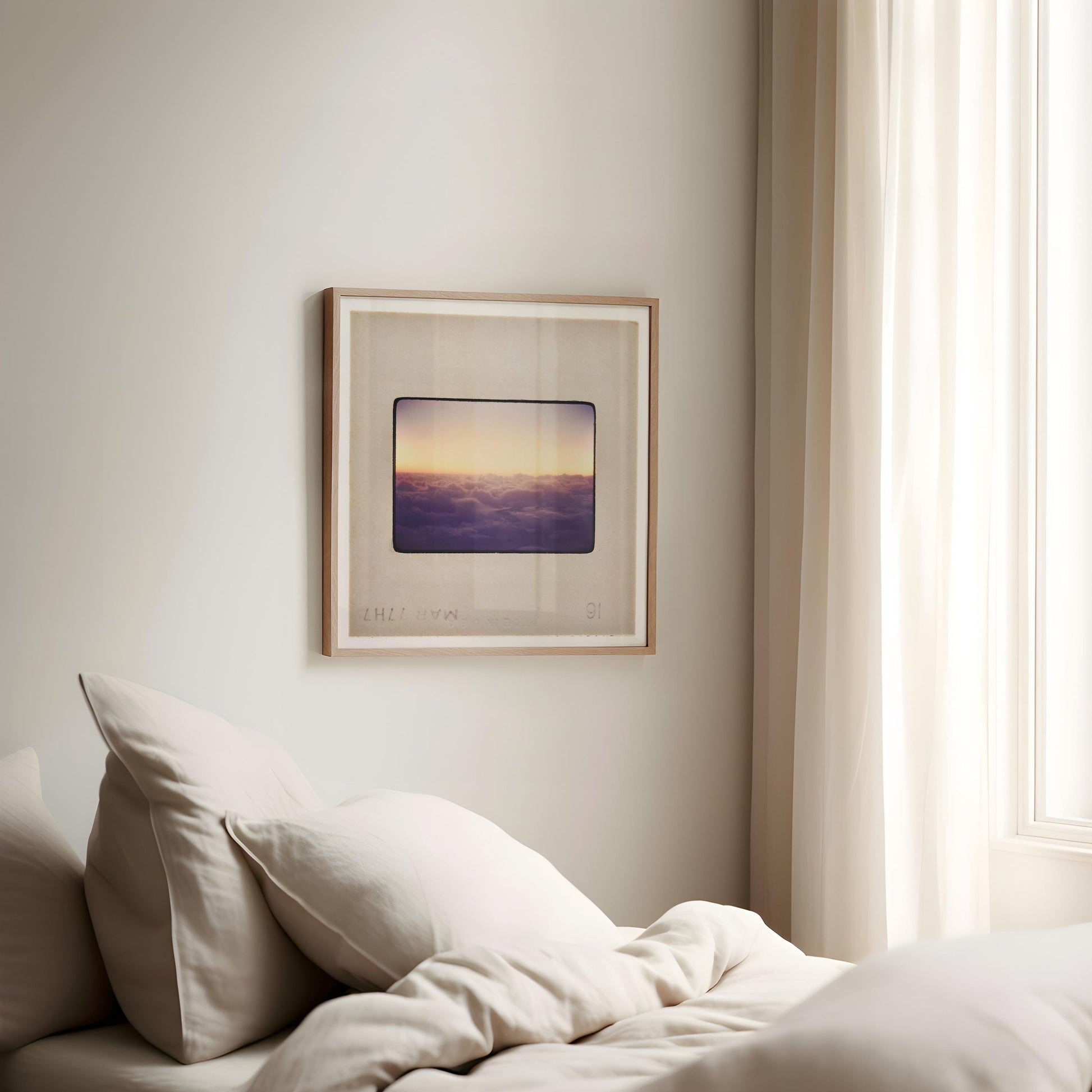 a cozy bedroom corner with a bed covered in white sheets and pillows, a framed picture of a sunset on the wall, and a window with sheer curtains.