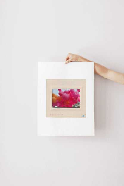 A person's hand is holding a white piece of paper with a framed image of colorful balloons on it.