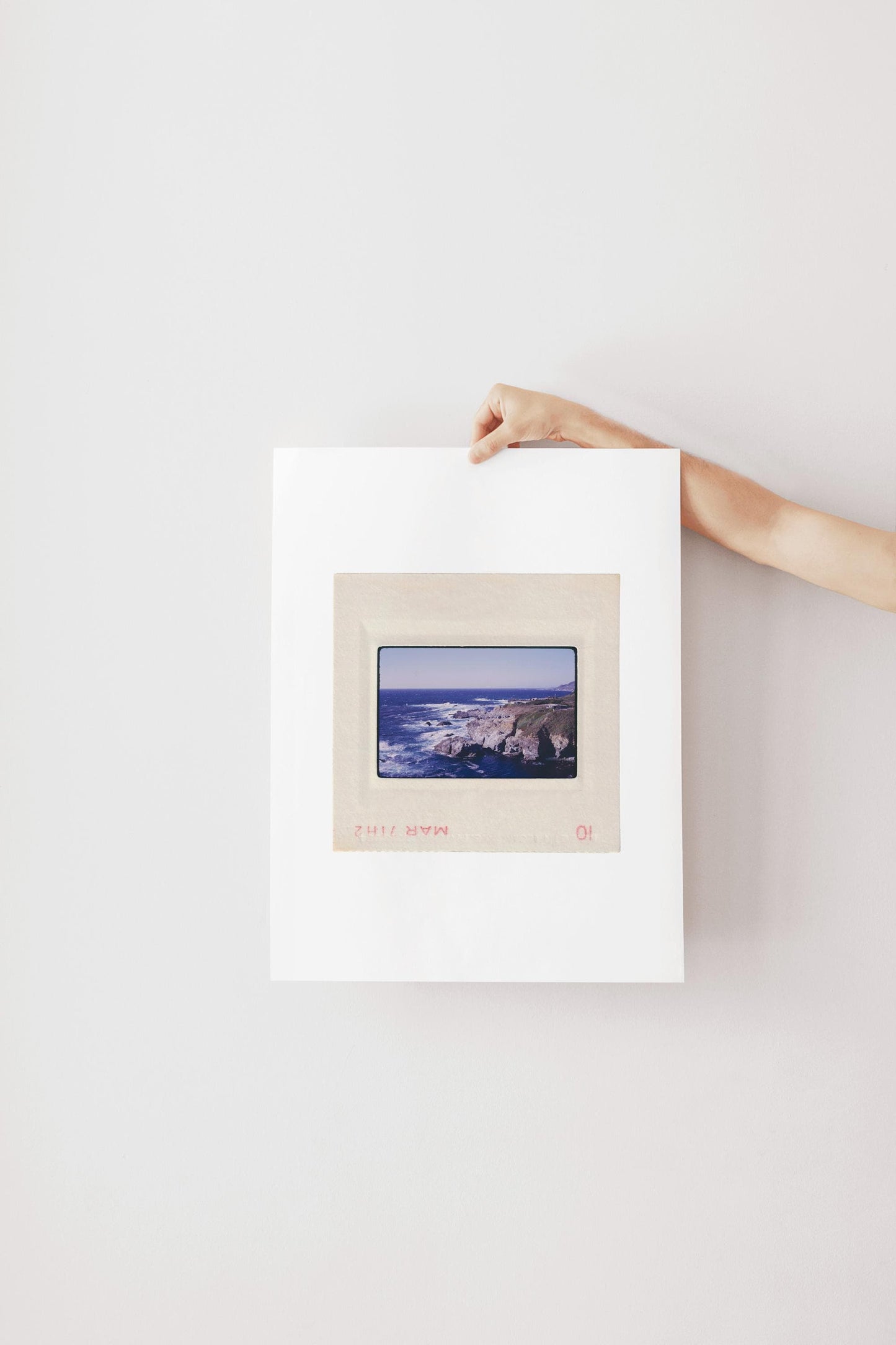 Vintage 1971 Kodachrome slide photograph of the Carmel California coastline, showing dramatic rocky cliffs and outcroppings meeting deep cobalt Pacific Ocean waters, with white surf, a green headland, and a small structure visible at the cliff's edge. Printed as a giclée fine art print on archival matte paper.