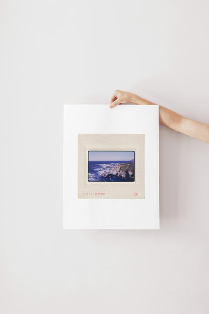 Vintage 1971 Kodachrome slide photograph of the Carmel California coastline, showing dramatic rocky cliffs and outcroppings meeting deep cobalt Pacific Ocean waters, with white surf, a green headland, and a small structure visible at the cliff's edge. Printed as a giclée fine art print on archival matte paper.