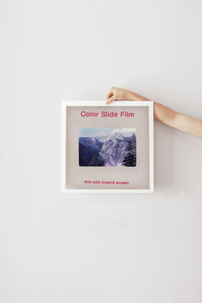 A person's hand is holding a white frame with a photograph of a mountain landscape inside it.
