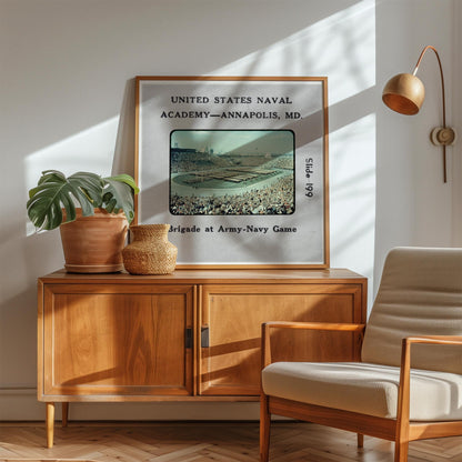 a framed poster of a baseball stadium, with text that reads "UNITED STATES NAVAL ACADEMY - ANNAPOLIS, MD" and "Slide 1969". The poster is displayed on a wooden cabinet, with a potted plant and a woven basket on top.