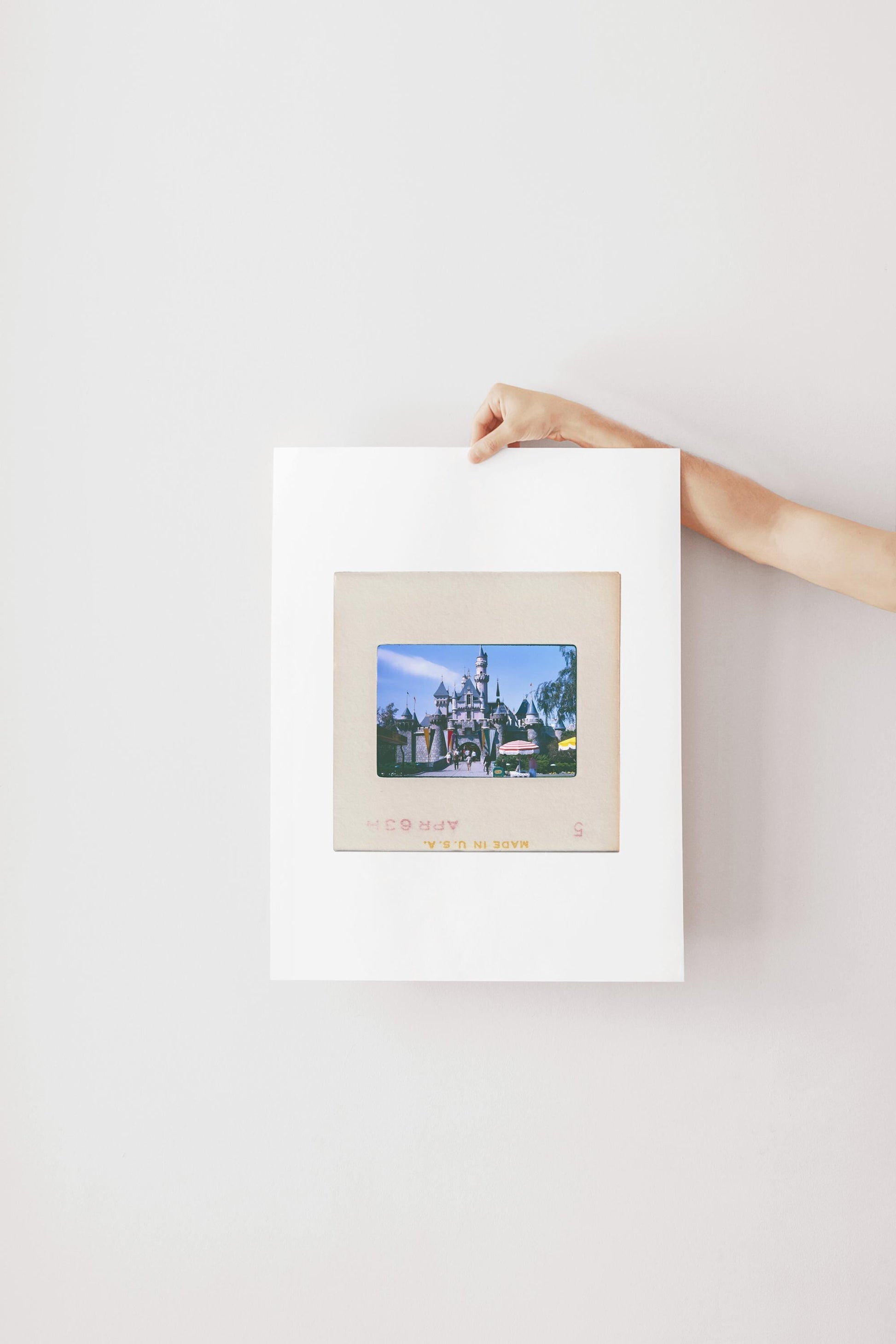 A person's hand is holding a framed picture of a castle or palace, which is hanging on a white wall.