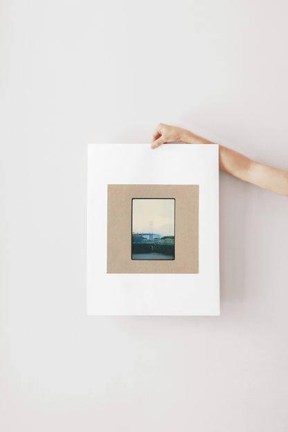 Vintage 35mm slide photograph of the Golden Gate Bridge partially obscured by dense fog, with only the tower visible above the cloud layer, viewed from a concrete overlook with green hedges in the foreground. Printed as a giclée fine art print on archival matte paper.