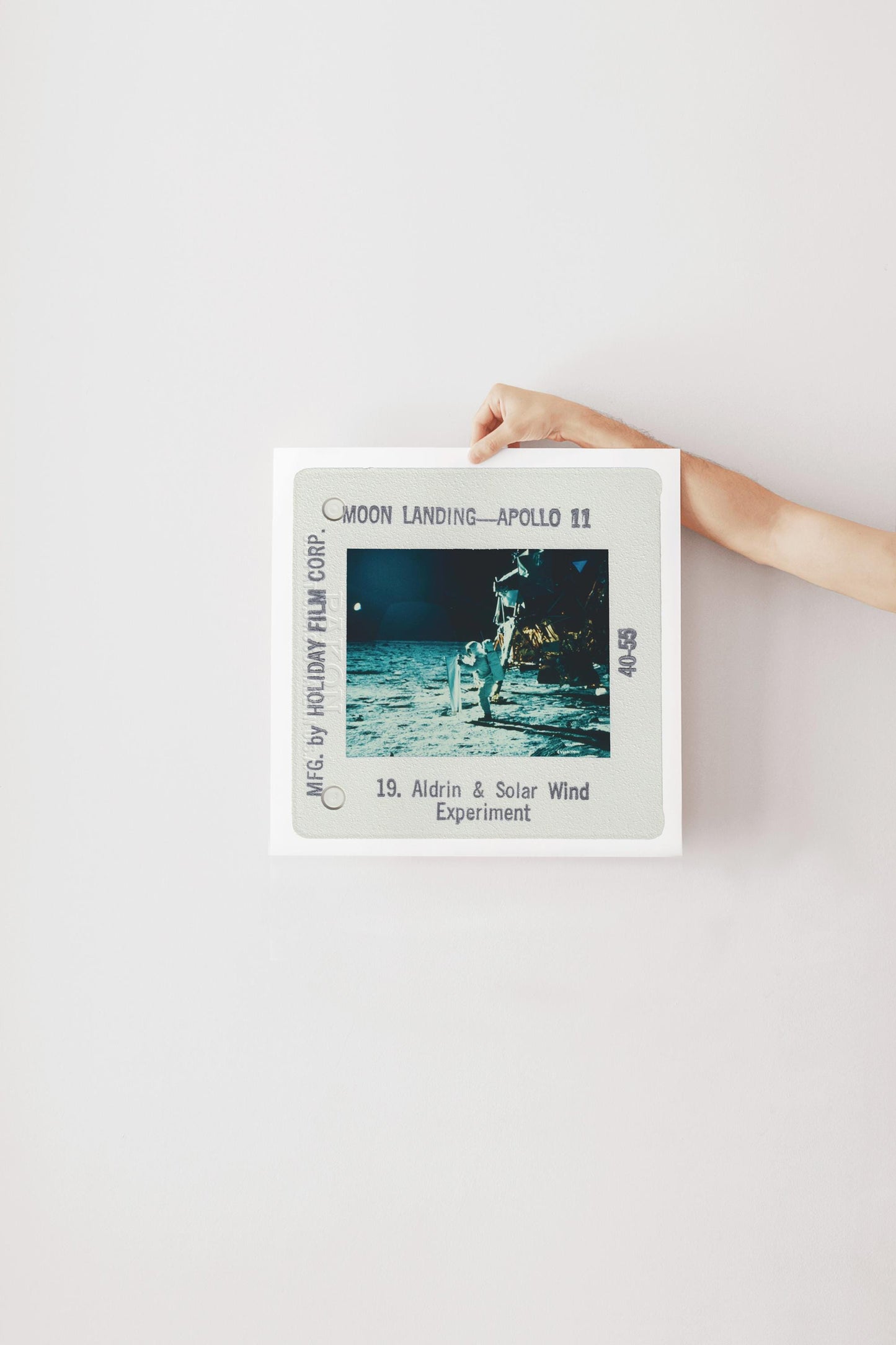 A person's hand is holding a framed photograph of the Apollo 11 moon landing, with the text "Moon Landing - Apollo 11" and "19. Aldrin & Solar Wind Experiment" visible.