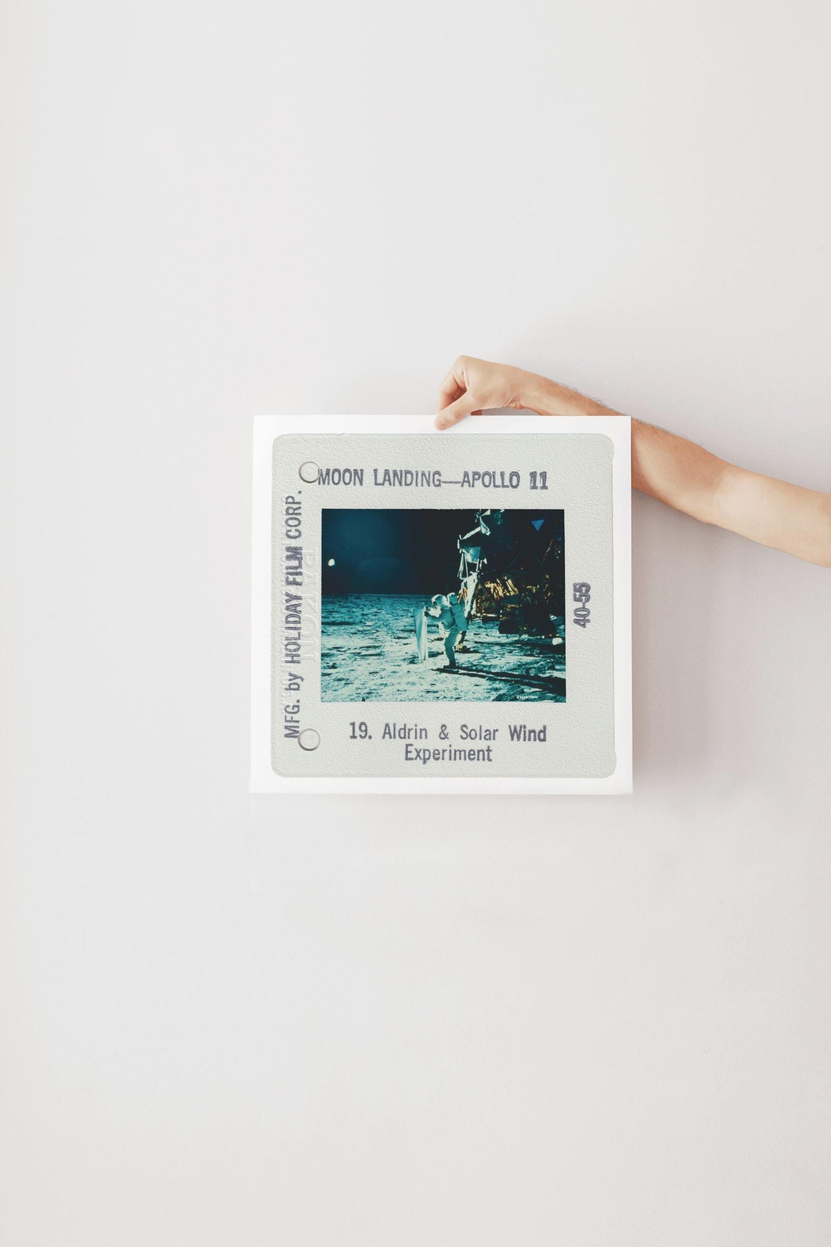 A person's hand is holding a framed photograph of the Apollo 11 moon landing, with the text "Moon Landing - Apollo 11" and "19. Aldrin & Solar Wind Experiment" visible.