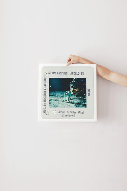 A person's hand is holding a framed photograph of the Apollo 11 moon landing, with the text "Moon Landing - Apollo 11" and "19. Aldrin & Solar Wind Experiment" visible.
