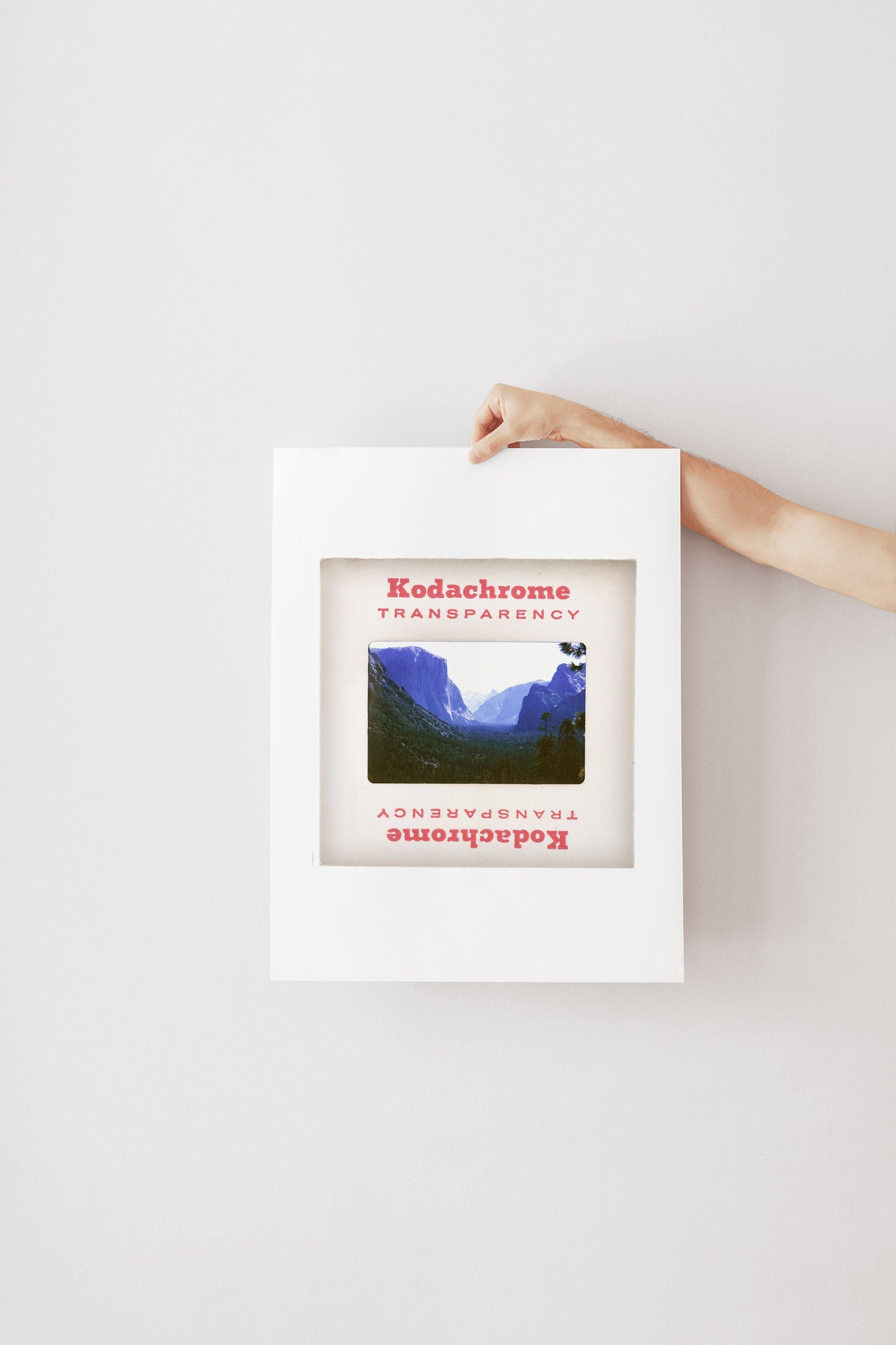 A hand is holding a framed photograph of a mountainous landscape, with the text "Kodachrome Transparency" visible on the frame.
