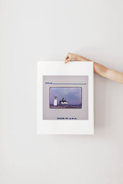 A person's hand is holding a framed photograph of a lighthouse against a white background.