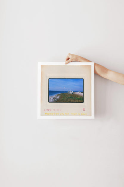 Vintage 1965 Kodachrome slide photograph of a canvas tent pitched on a grassy coastal dune in California, with a camper vehicle in the background, calm blue water, and a single seagull in flight overhead. Printed as a giclée fine art print on archival matte paper.