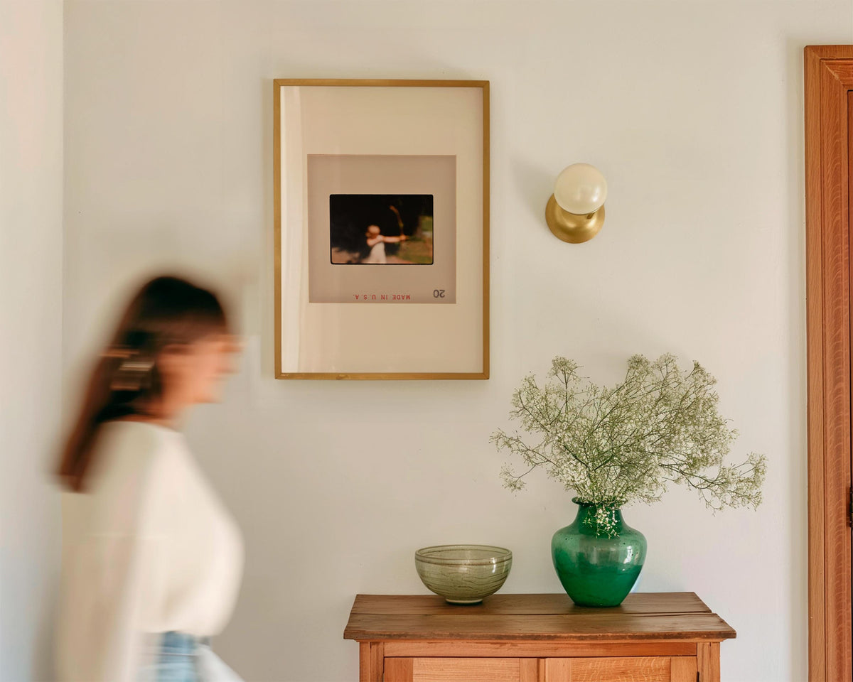 a woman walking past a wooden table with a framed photo, a green vase with flowers, and a bowl on it. The photo appears to be a portrait of a child.
