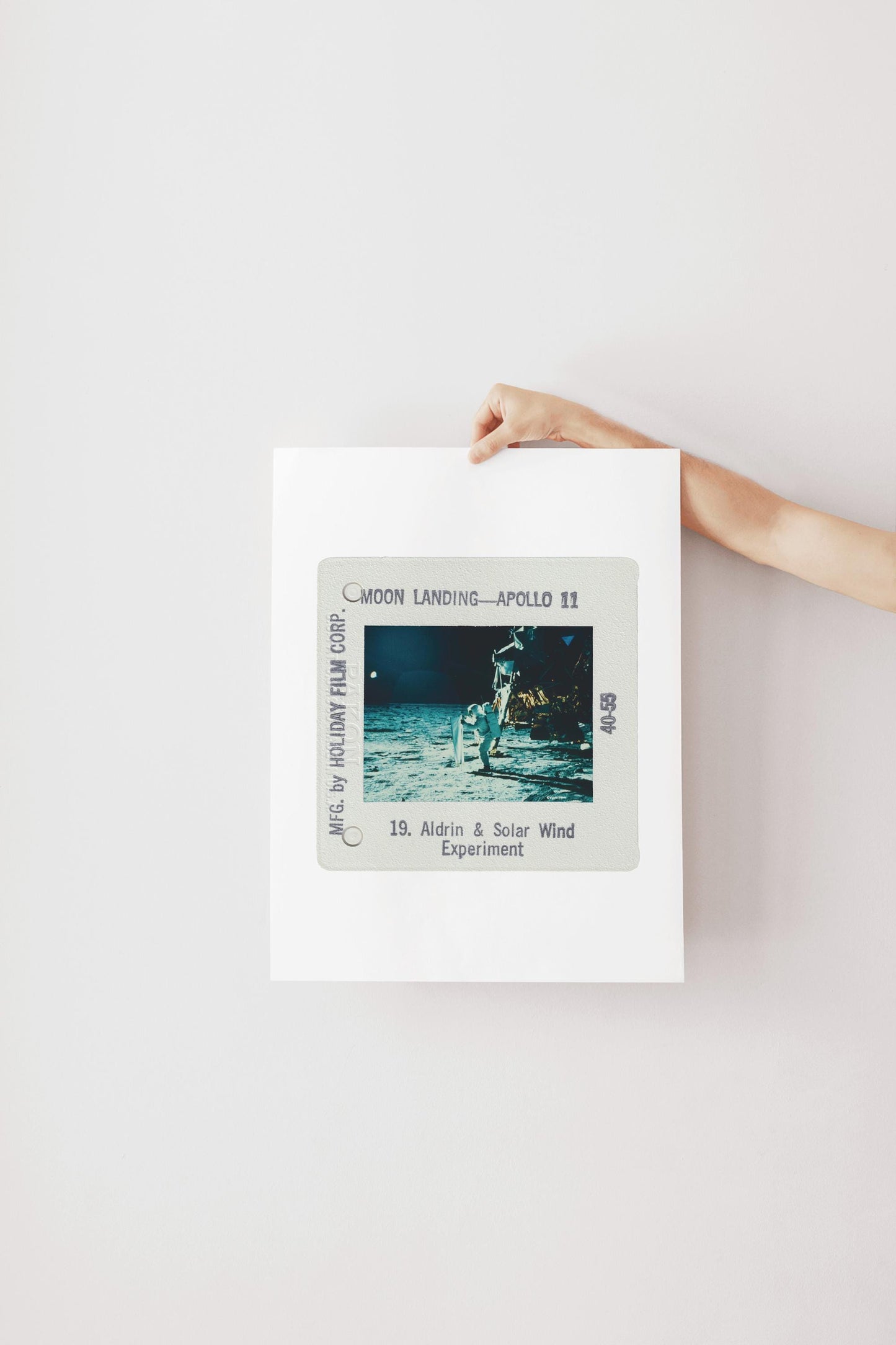 A person's hand is holding a framed photograph of the Apollo 11 moon landing, with the text "Moon Landing - Apollo 11" and "18. Aldrin & Solar Wind Experiment" visible.