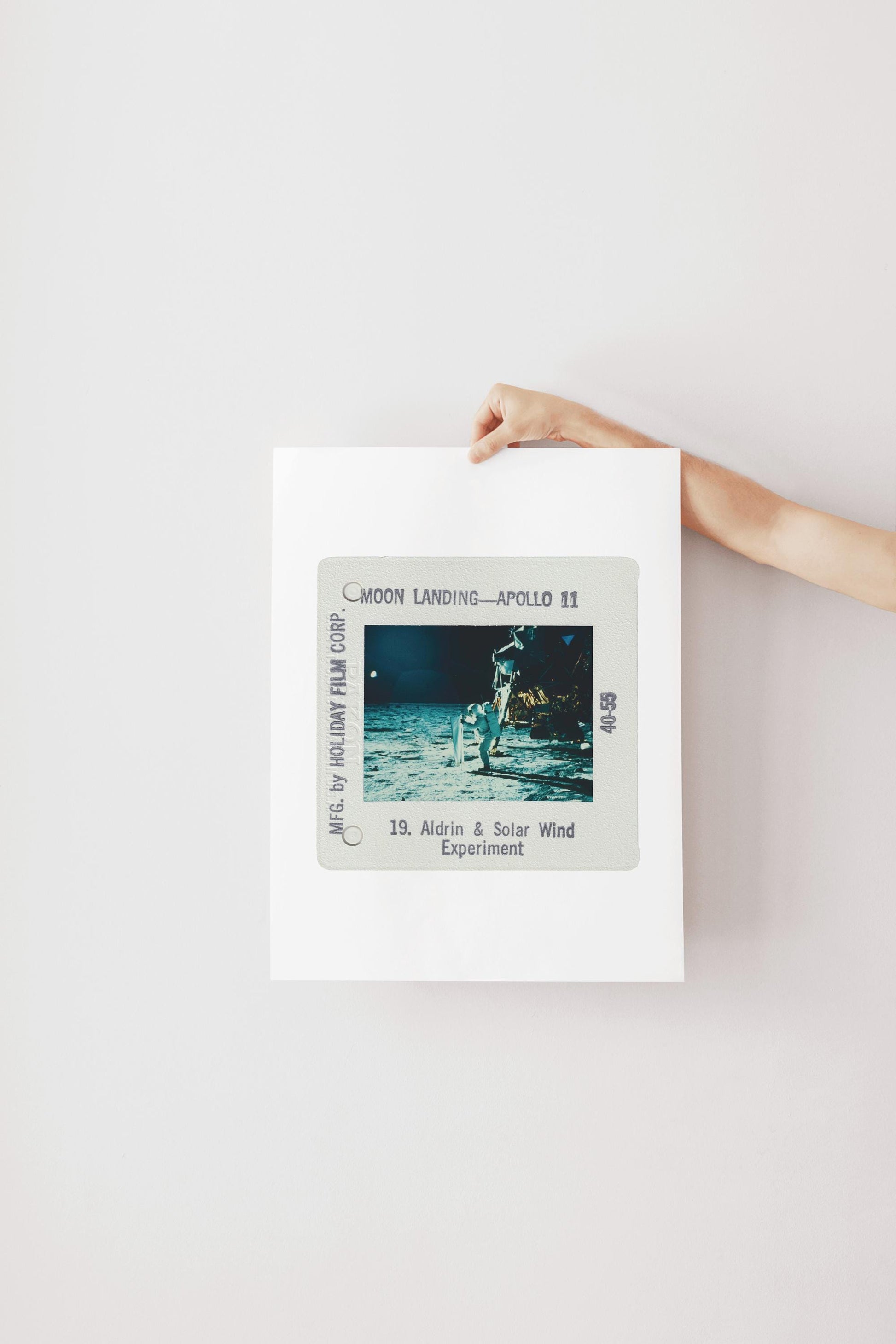 A person's hand is holding a framed photograph of the Apollo 11 moon landing, with the text "Moon Landing - Apollo 11" and "18. Aldrin & Solar Wind Experiment" visible.