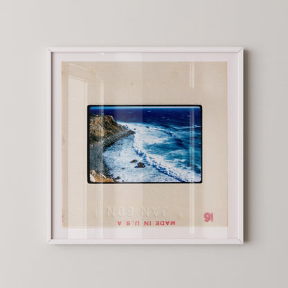 Vintage 1960 Kodachrome slide photograph taken from an elevated vantage point overlooking a rugged California rocky coastline, with ochre-colored cliffs curving into the frame, white surf breaking against dark rocks, deep cobalt blue Pacific Ocean, and a small structure visible at the cliff's edge. Printed as a giclée fine art print on archival matte paper.